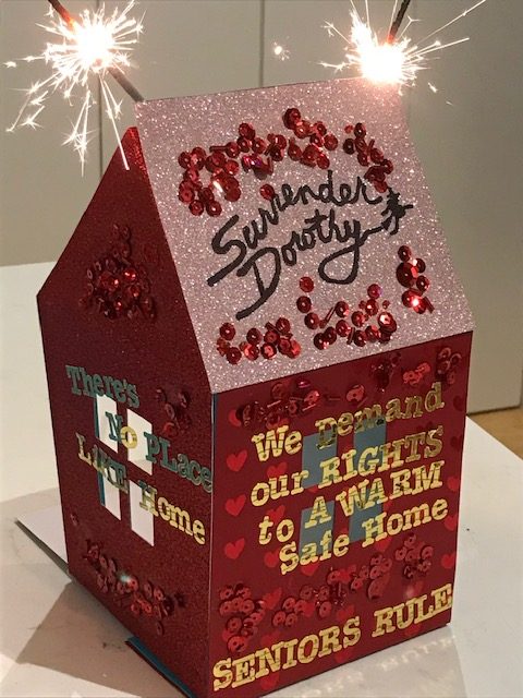 Cardboard house decorated in red glitter and the words 'We demand our right to a warm safe home'