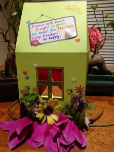 A green warm safe home made of cardboard decorated with flowers and the words 'A warm safe home. The right to grow, be cared for, feel safe, enjoy freedom, be happy.'